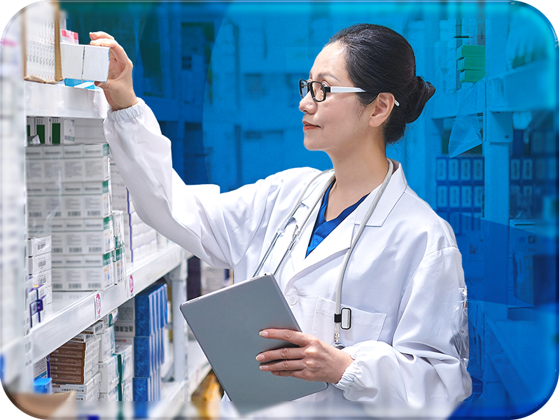 A female medical professional in a white lab coat and stethoscope stands in a medical supply room. She reaches with her right hand toward a shelf filled with various boxes of medical supplies while holding a gray tablet in her left hand. She wears black-framed glasses, and her dark hair is pulled back. The background features rows of shelves stocked with colorful boxes, slightly blurred, with a blue overlay adding a modern, digital feel that draws attention to the healthcare professional.