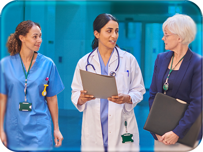 Three professional women walk through a bright hospital corridor, engaged in conversation. On the left, a woman with curly dark hair wears light blue scrubs, a green lanyard, and a yellow device clipped to her top. In the center, a doctor with dark hair tied back wears a white lab coat over blue scrubs, a stethoscope around her neck, and holds a gray folder. On the right, a woman with short white hair and glasses wears a dark blue blazer over a light top and skirt, carrying a dark folder under her arm. The background shows a clean, well-lit hospital hallway with light-colored walls and reflective floors, creating a professional and collaborative atmosphere.