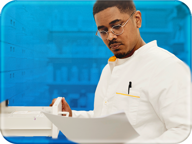 A male professional, likely a scientist or healthcare worker, is shown in a white lab coat with orange-yellow trim, holding papers in his right hand and a white device in his left. He has short dark hair, a neatly trimmed beard, and wears round gold-rimmed glasses, looking down intently at the papers. The background is a soft blue with blurred hints of shelves and healthcare equipment, suggesting a laboratory or clinical setting. The image conveys focus and professionalism. 