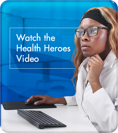 A young, professional woman with long, wavy, brown-colored hair, wearing glasses and a white lab coat, sits at a desk in a professional healthcare setting. She is focused on a computer. There is wording on the image reading: Watch the Health Heros Video.