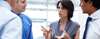 A woman in business attire with a few other people surrounding her, gestures with her hands during a conversation